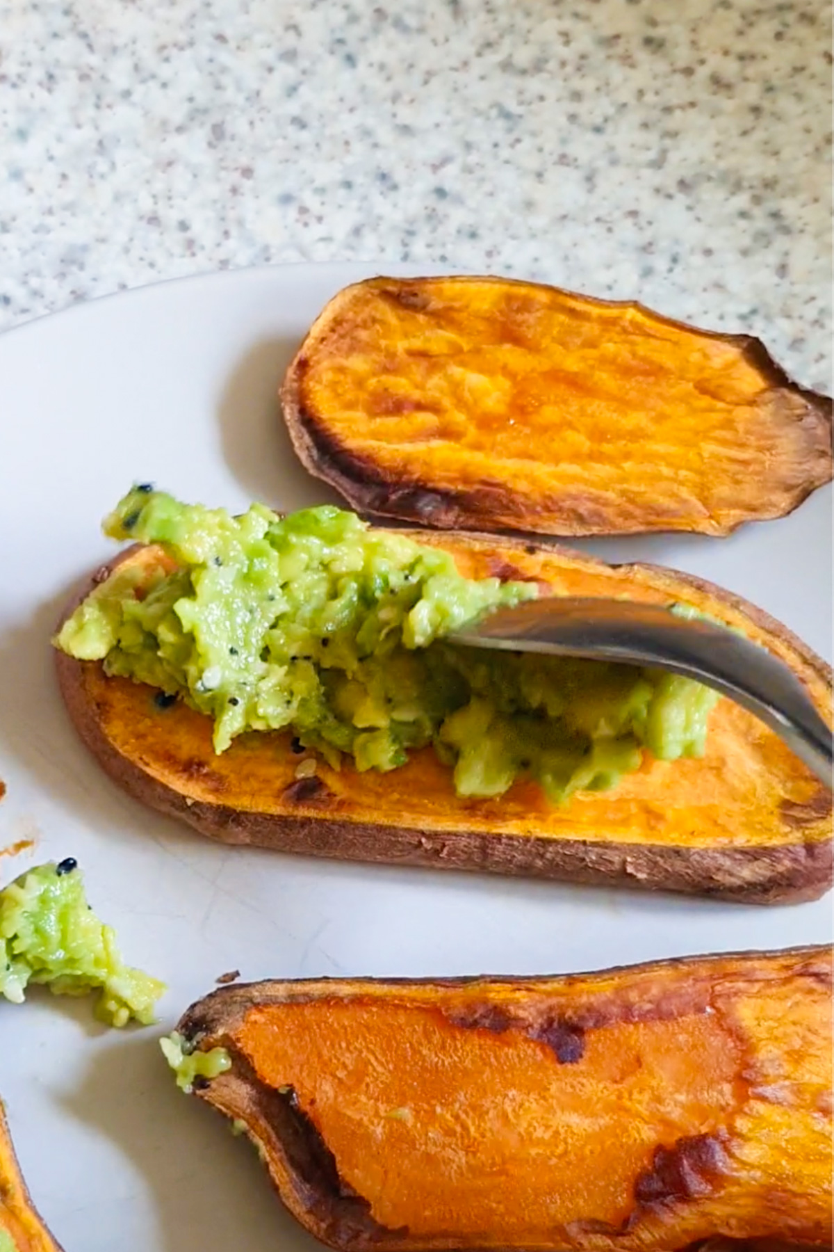 Sweet potato toast on a white plate, being topped with mashed avocado mix by a spoon.