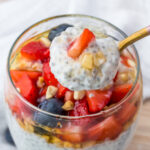 A glass filled with yogurt chia seed pudding, topped with chopped strawberries, blueberries, and nuts. A spoon holds a scoop of the pudding above the glass.