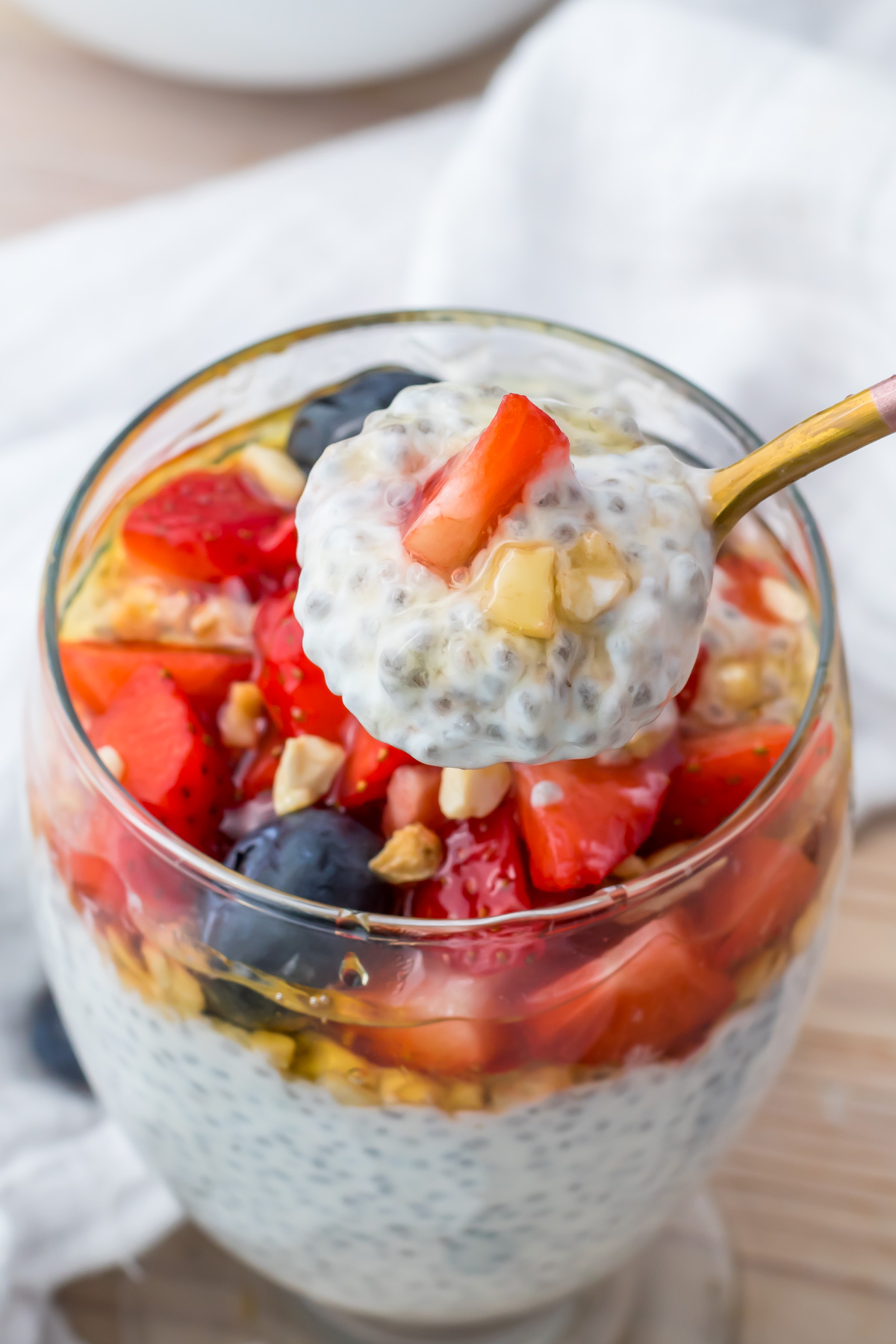 A glass filled with yogurt chia seed pudding, topped with chopped strawberries, blueberries, and nuts. A spoon holds a scoop of the pudding above the glass.