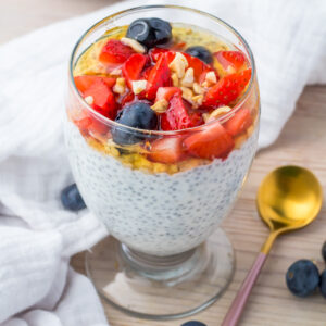 A glass of yogurt chia pudding topped with strawberries, blueberries, granola, and honey sits on a table next to a gold spoon and a white napkin.