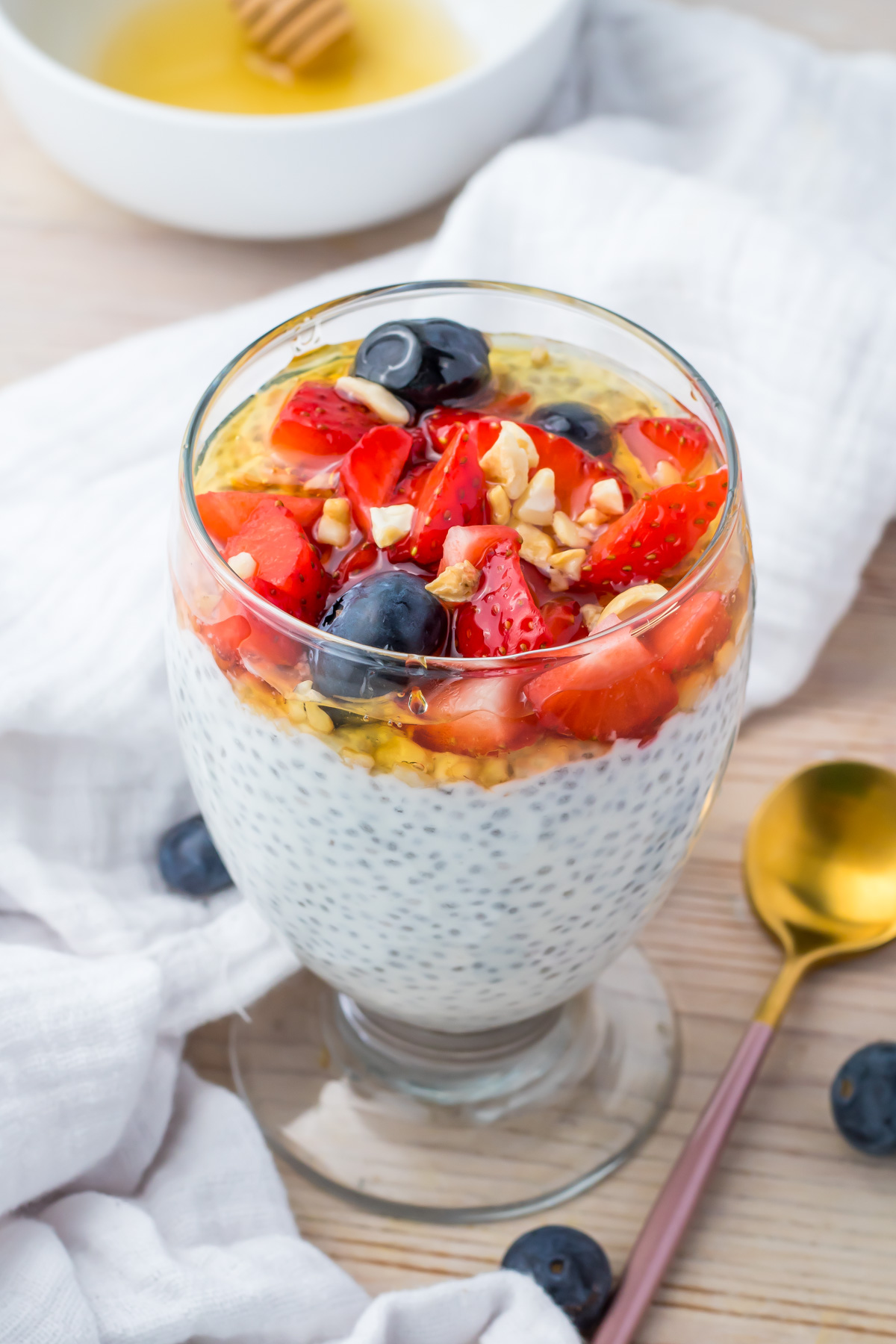 A glass filled with chia seed pudding topped with chopped strawberries, blueberries, walnuts, and a drizzle of honey. A gold spoon and bowl of honey are nearby.