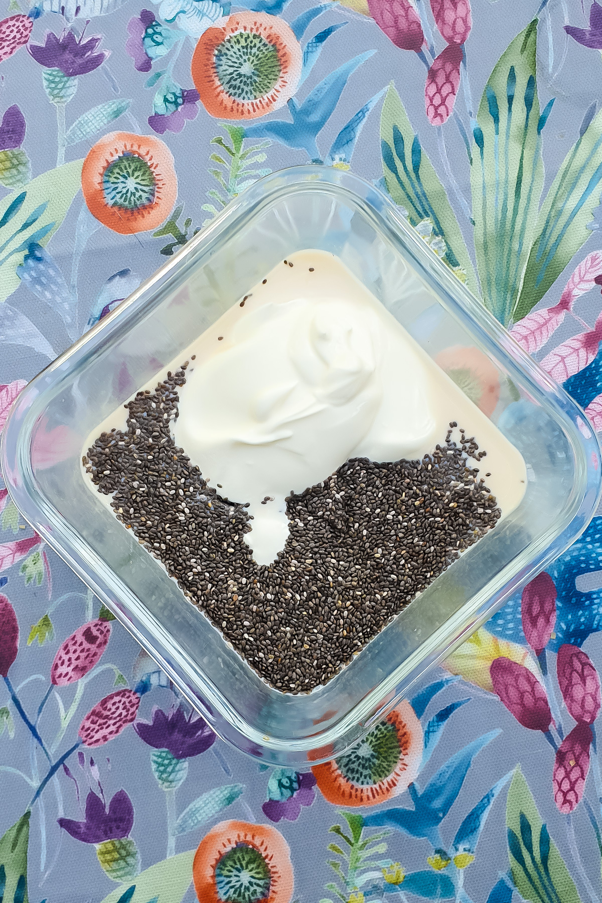 A glass container filled with plain yogurt, chia seeds, and milk, placed on a colorful floral-patterned surface.
