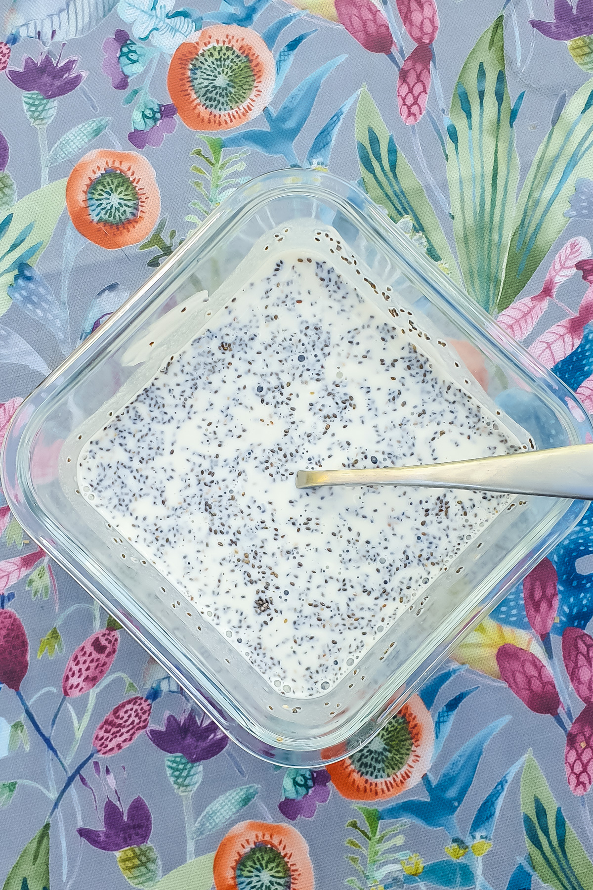 A square plastic container filled with chia seeds mixed in milk and yogurt sits on a colorful, floral-patterned surface. A spoon rests inside the mixture.
