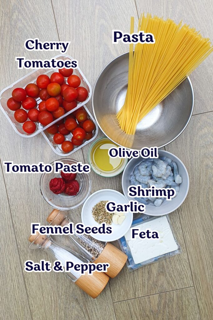 Ingredients for a shrimp feta pasta dish laid out: uncooked spaghetti, cherry tomatoes, shrimp, olive oil, tomato paste, garlic, feta, fennel seeds, salt, and pepper on a wood surface.
