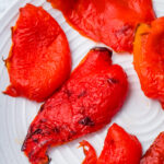 Several pieces of roasted red bell pepper with slightly charred skins are arranged on a white plate.