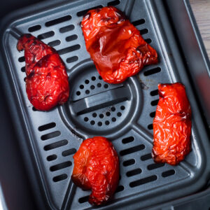 Four air fryer roasted red bell peppers with wrinkled skins are arranged inside an air fryer basket.