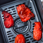 Four roasted red bell pepper halves sit inside an air fryer basket, with charred and wrinkled skins visible.