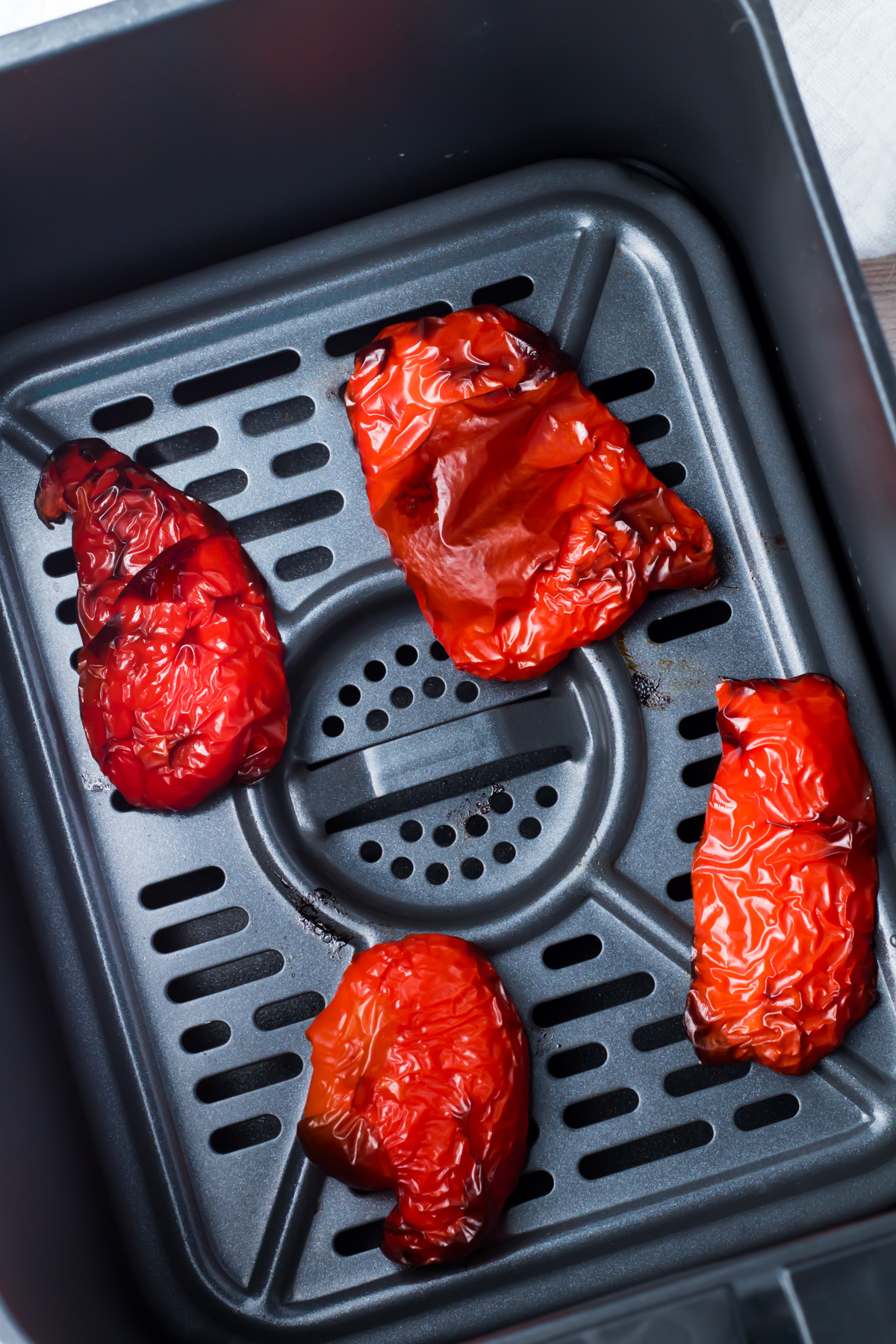 Four roasted red bell pepper halves sit inside an air fryer basket, with charred and wrinkled skins visible.