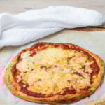 A cottage cheese pizza with a golden crust sits on parchment paper next to a pizza cutter and a white cloth on a wooden surface.