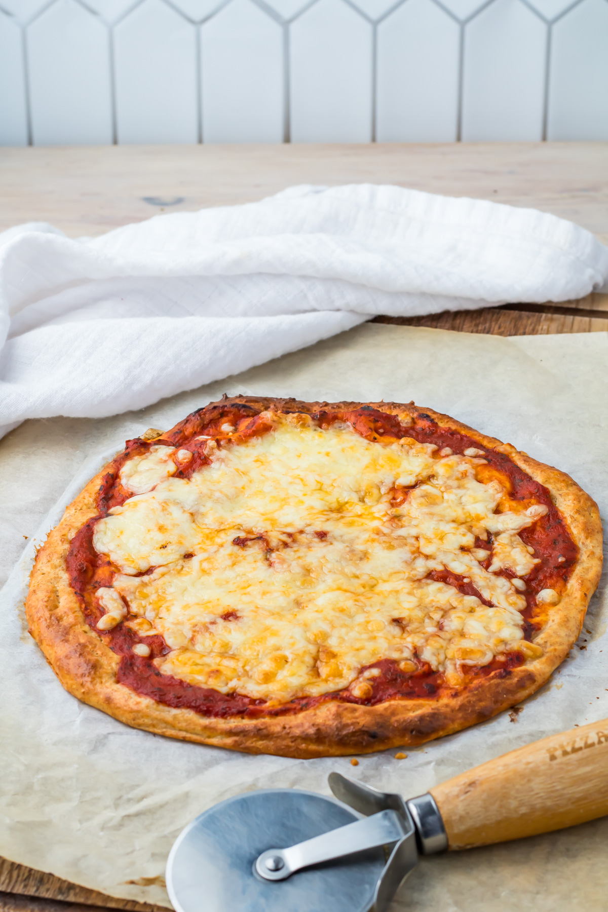 A cottage cheese pizza with a golden crust sits on parchment paper next to a pizza cutter and a white cloth on a wooden surface.
