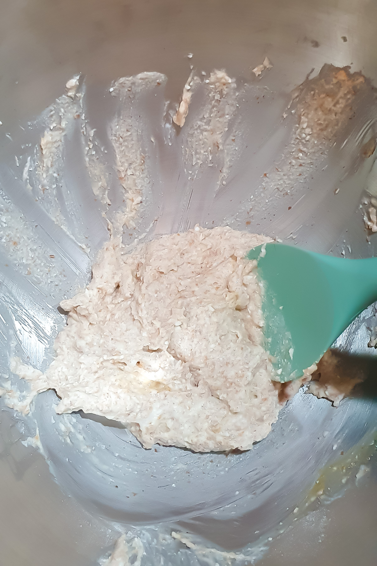 A teal spatula mixing a thick, pale batter in a large stainless steel bowl. Some batter is spread along the sides of the bowl.