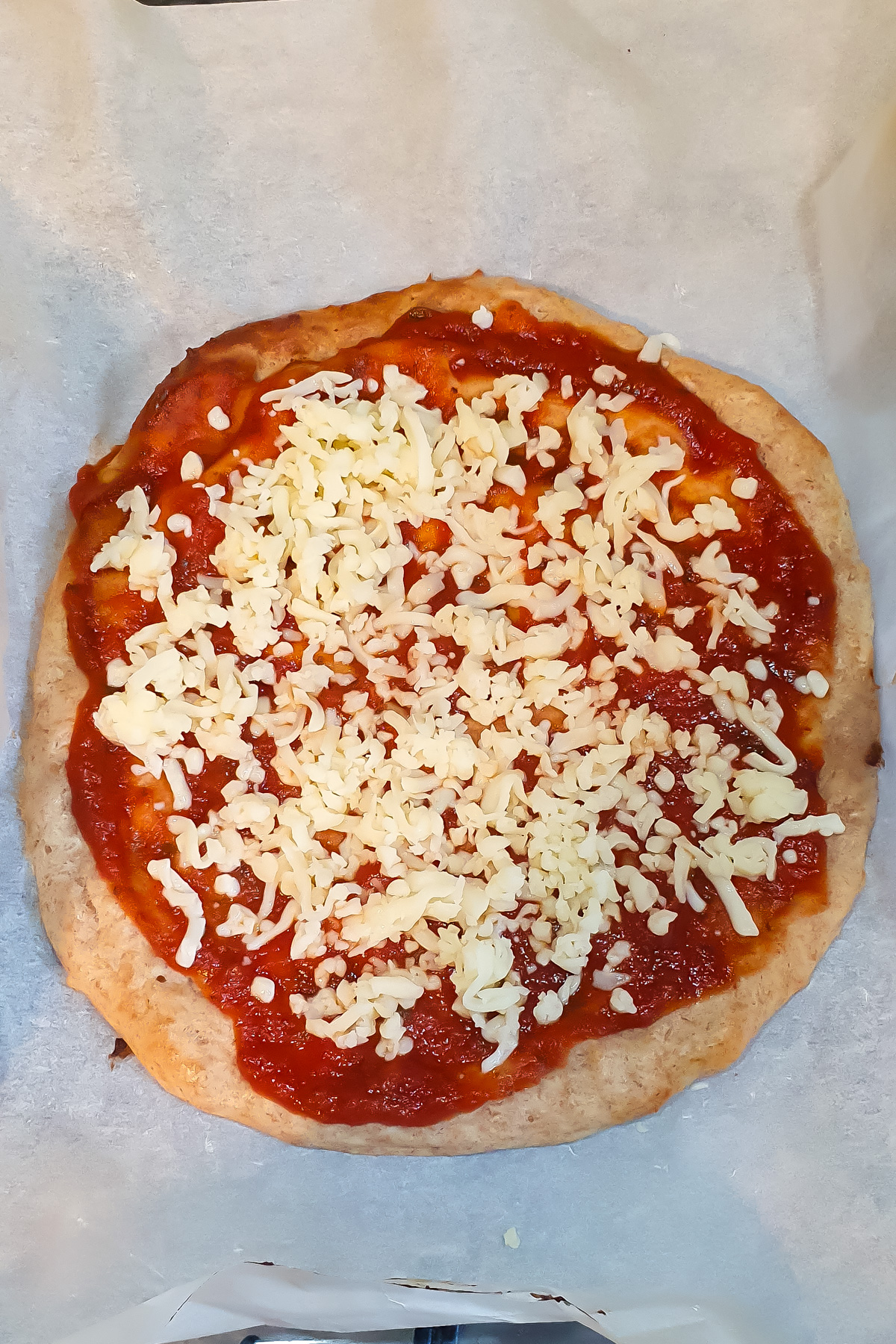 A round pizza crust topped with tomato sauce and shredded mozzarella cheese on a sheet of parchment paper.