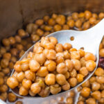 A close-up of a slotted spoon lifting cooked chickpeas from an instant pot with liquid.
