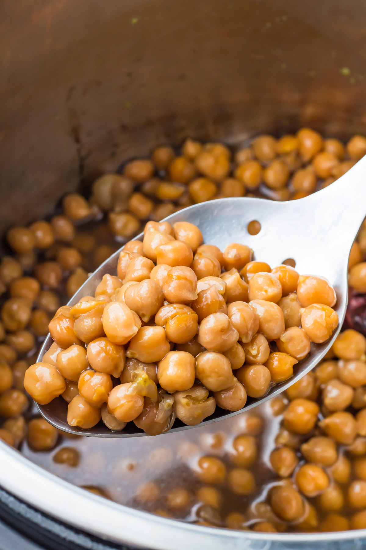 A close-up of a slotted spoon lifting cooked chickpeas from an instant pot with liquid.