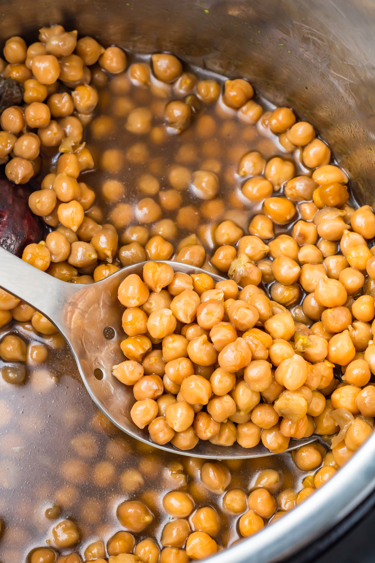 A metal spoon scooping cooked instant pot chickpeas from a pot with brown cooking liquid.