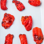 Eight roasted red peppers with wrinkled, blistered skin are arranged on a white surface.