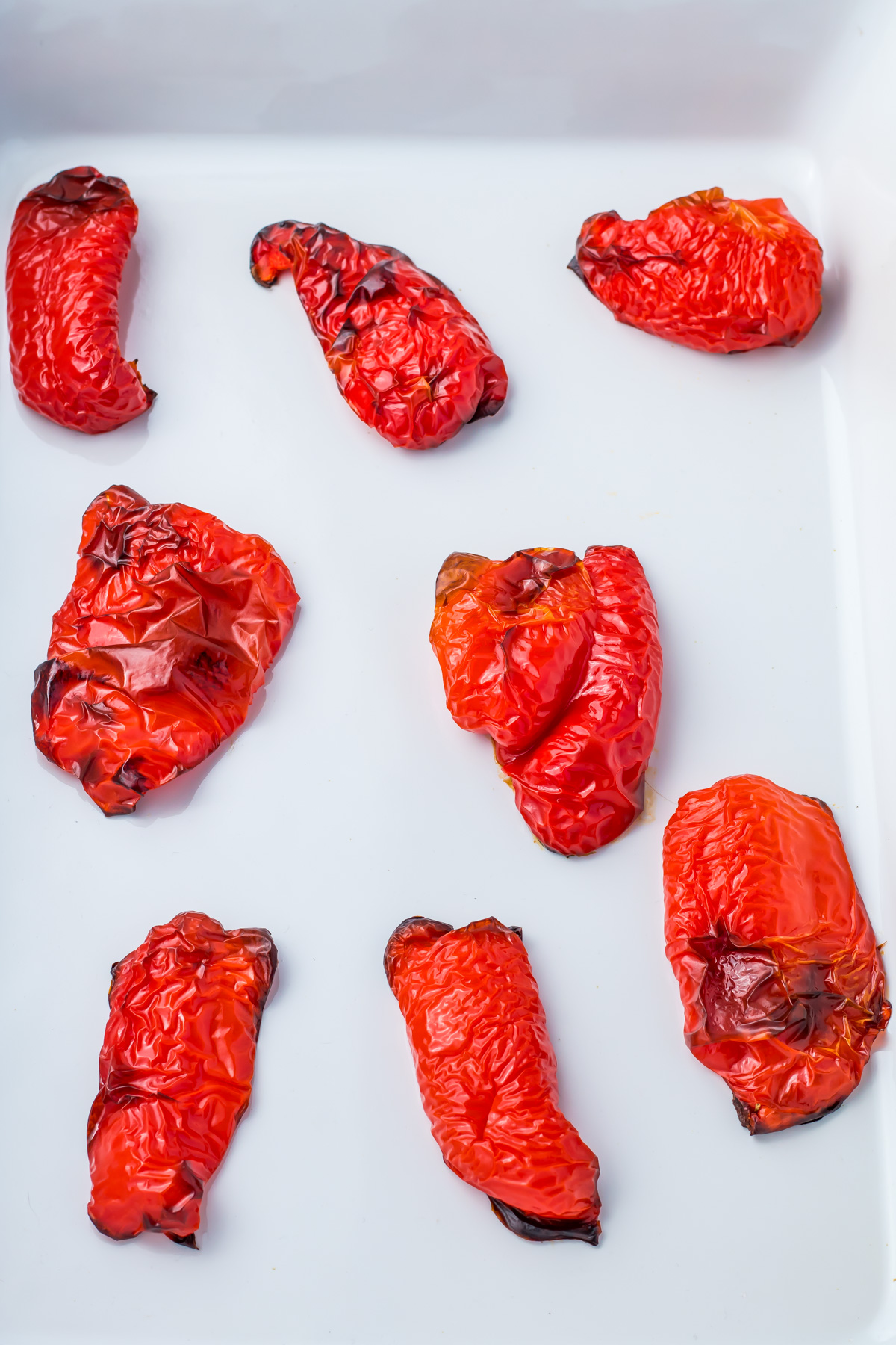 Eight roasted red peppers with wrinkled, blistered skin are arranged on a white surface.