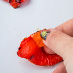 A hand peels the skin off a roasted red bell pepper on a white surface.