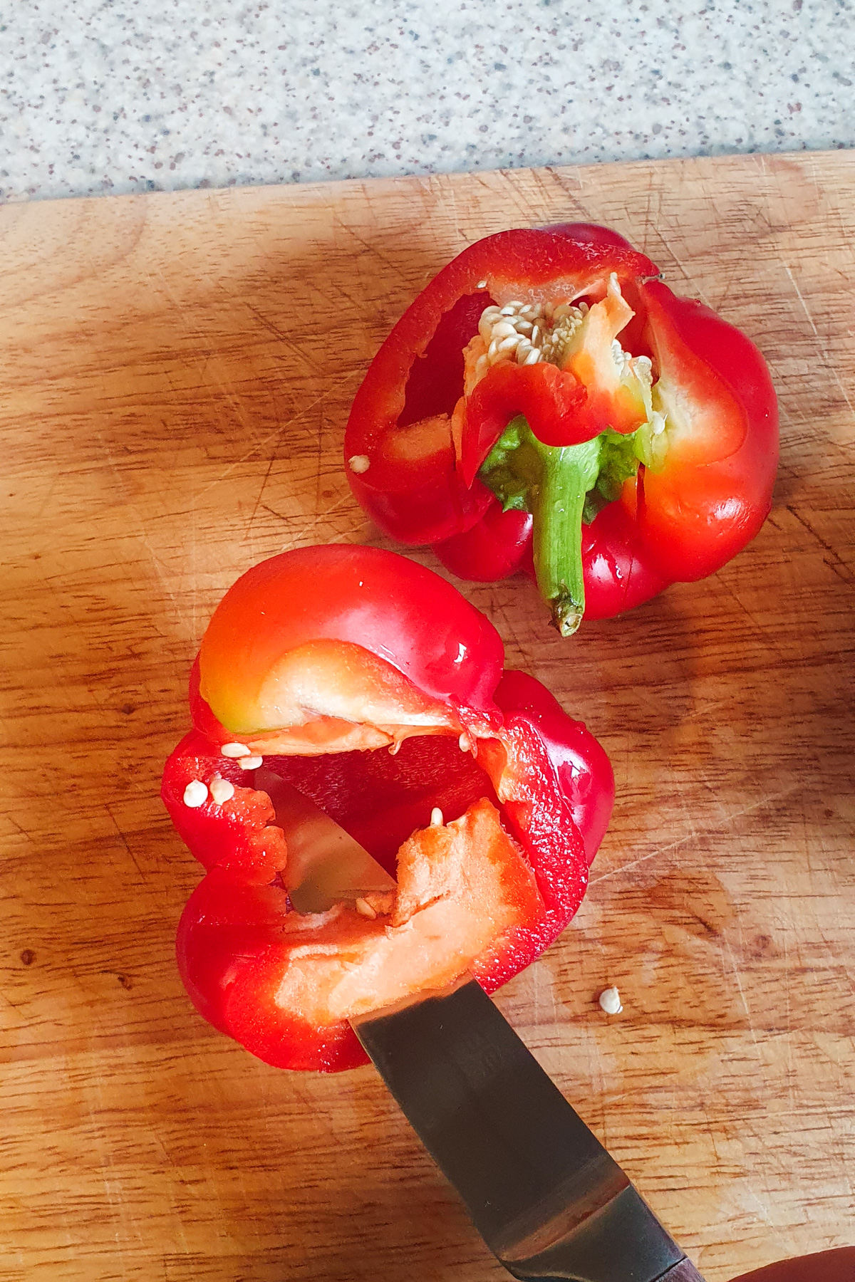 A red bell pepper cut open on a wooden cutting board, with seeds visible and a knife inserted into one half.