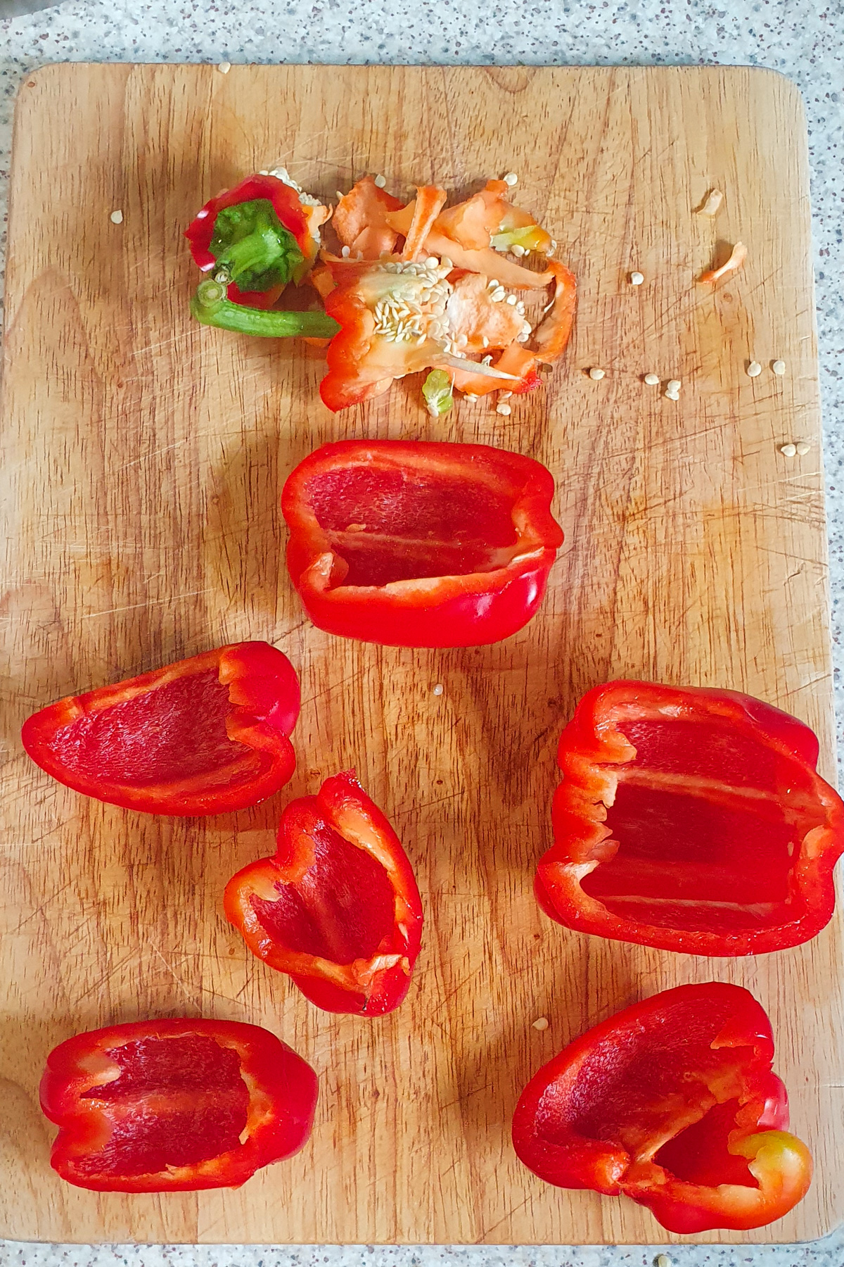 Six red bell pepper halves and pepper stems with seeds are on a wooden cutting board, placed on a speckled countertop.