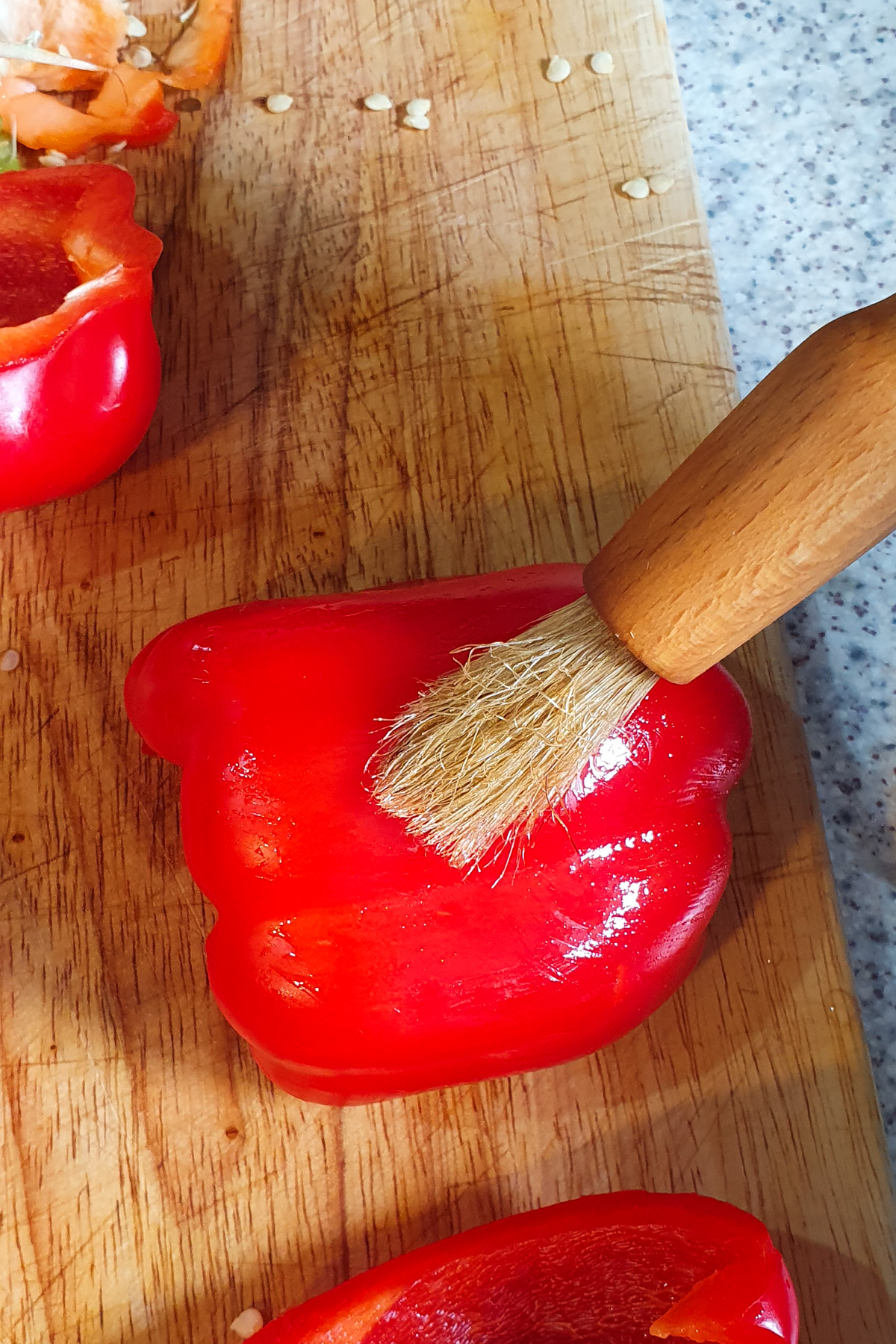 A wooden brush applies oil to a red bell pepper on a wooden cutting board, with chopped pepper pieces and seeds nearby.