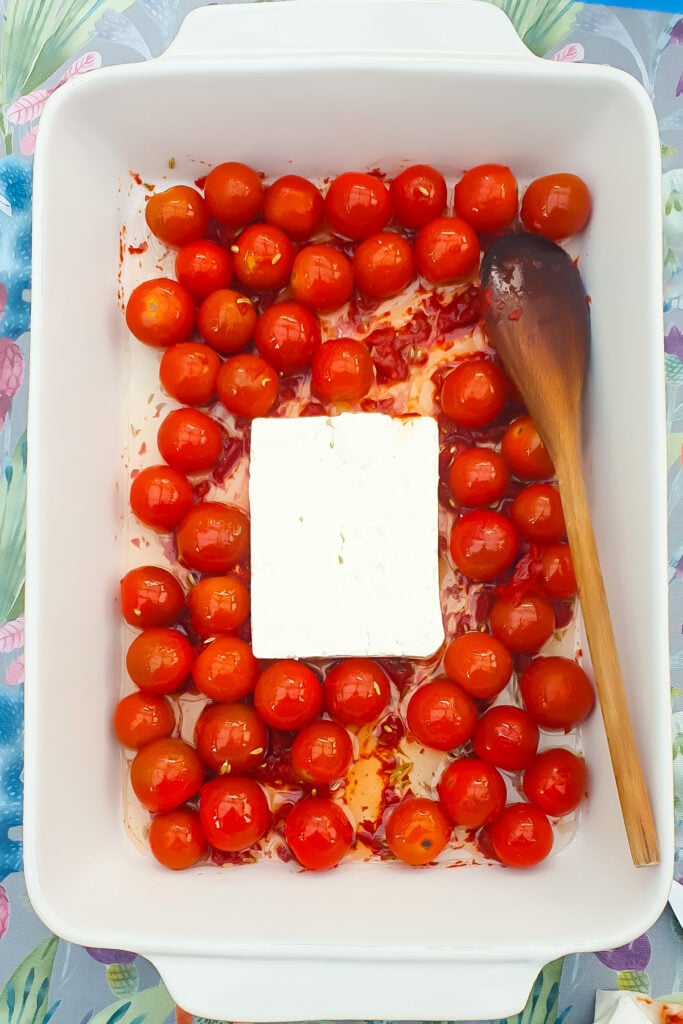 A white baking dish with cherry tomatoes, a block of feta cheese in the center, and a wooden spoon on the right side.