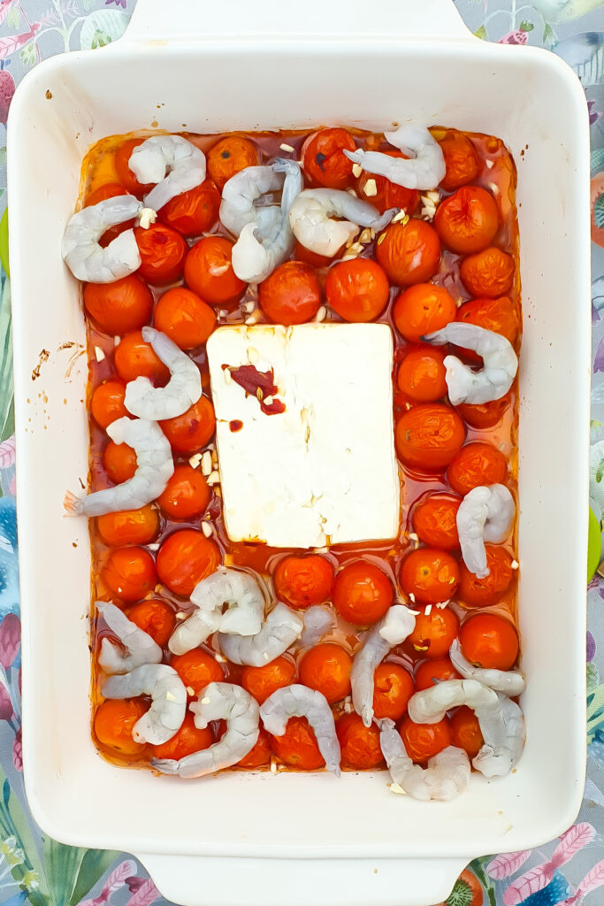 Raw shrimp, whole cherry tomatoes, minced garlic, and a block of feta cheese are arranged in a white baking dish, ready to be cooked.