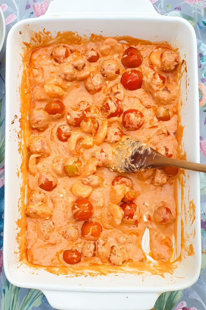 A white baking dish filled with cooked shrimp, cherry tomatoes, and a creamy orange sauce, with a wooden spoon resting inside.