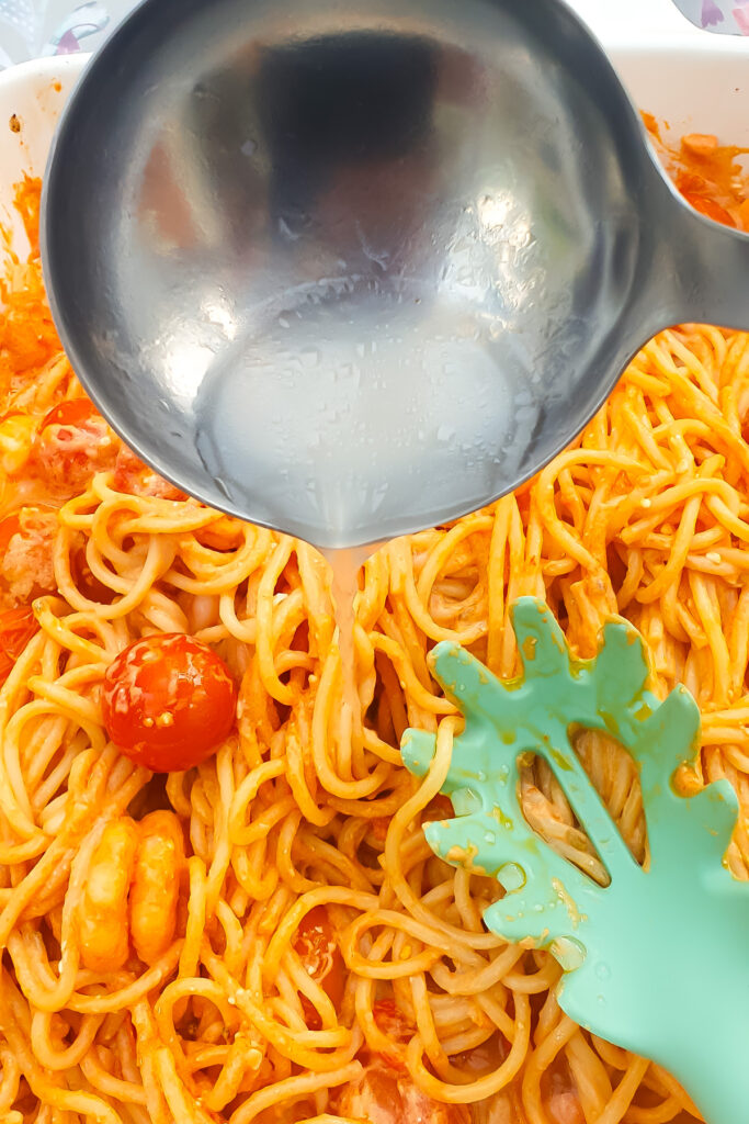 A metal ladle pours pasta water onto a dish of cooked spaghetti with tomato sauce; a green pasta fork rests on the noodles.
