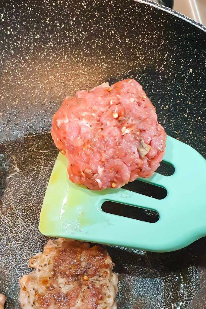 A raw turkey sausage patty is held on a green spatula above a frying pan, with a cooked patty visible beside it.