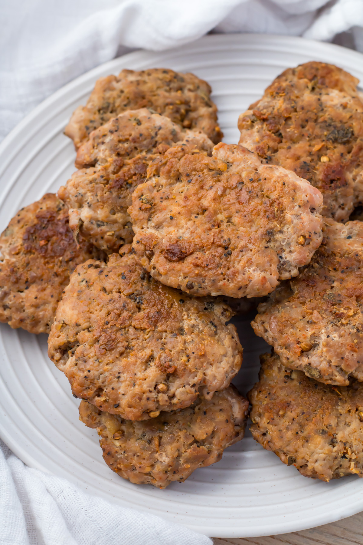 A plate with several cooked turkey sausage patties stacked on top of each other, placed on a white surface with a white cloth in the background.