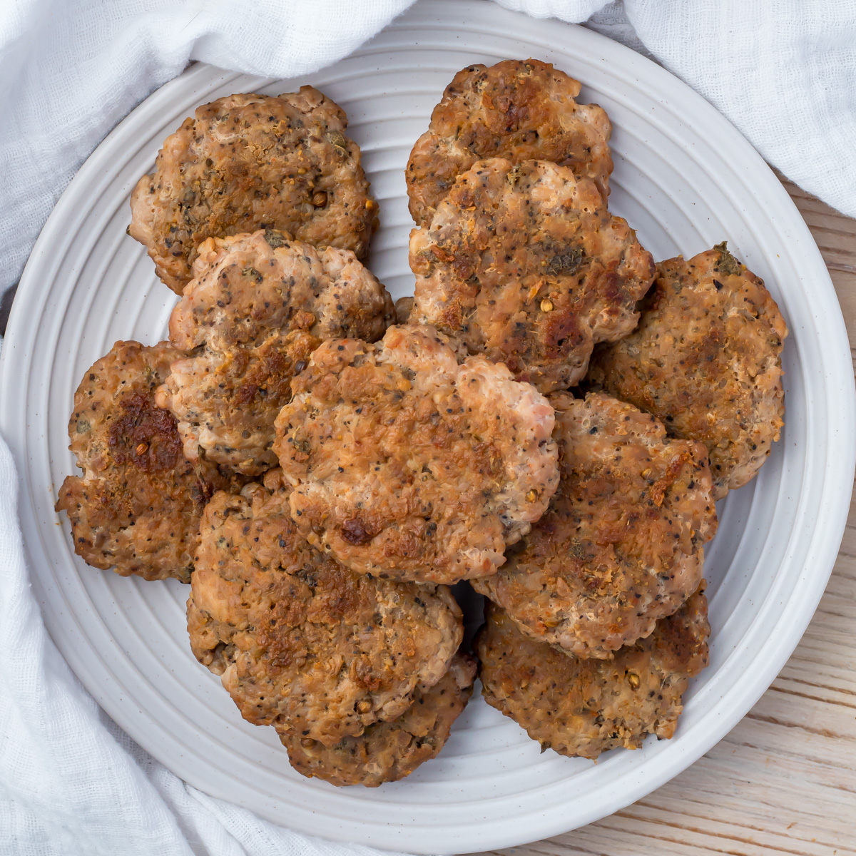 A white plate filled with several browned, seasoned turkey sausage patties, set on a light wooden surface with a white cloth in the background.