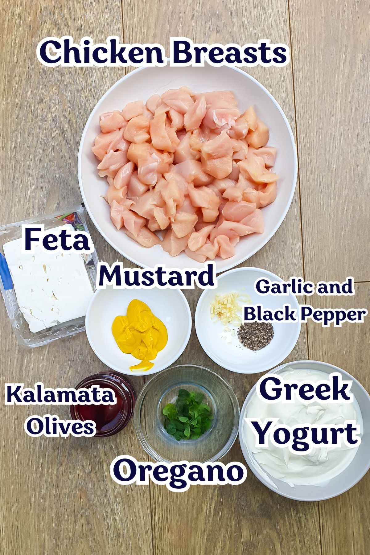 A bowl of diced chicken breasts surrounded by labeled ingredients: feta, mustard, garlic and black pepper, Kalamata olives, oregano, and Greek yogurt on a wooden surface.