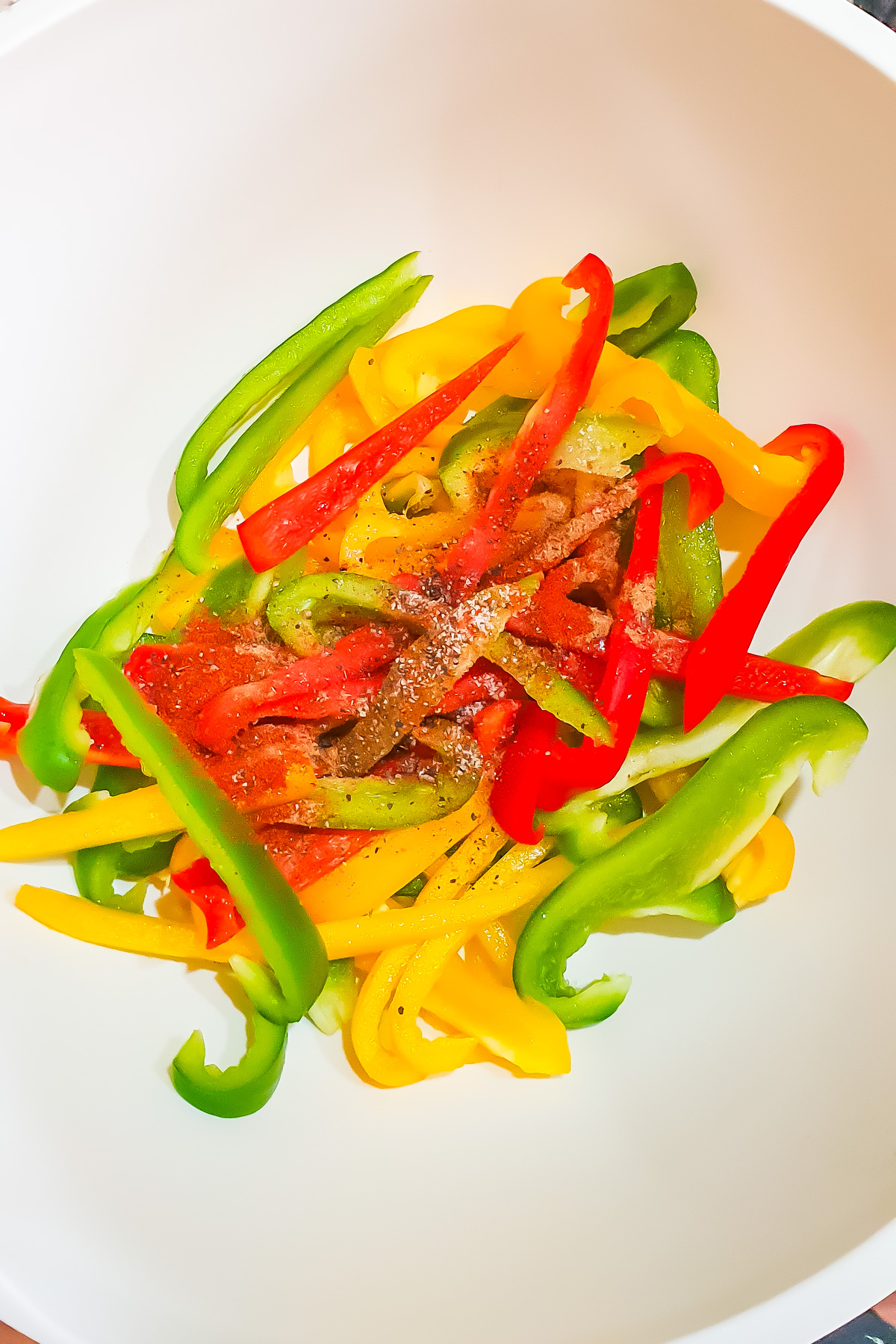 Sliced green, yellow, and red bell peppers with seasoning in a white bowl.