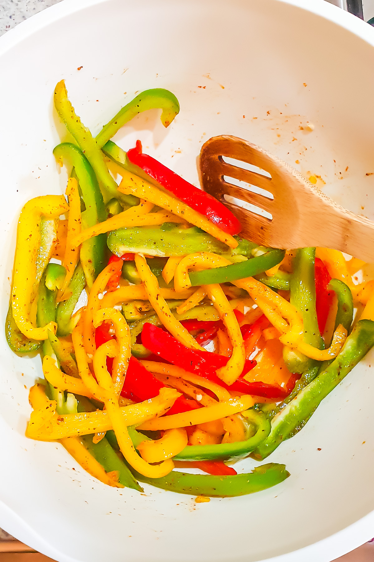 A white bowl containing sliced yellow, red, and green bell peppers seasoned with spices, with a wooden slotted spoon.
