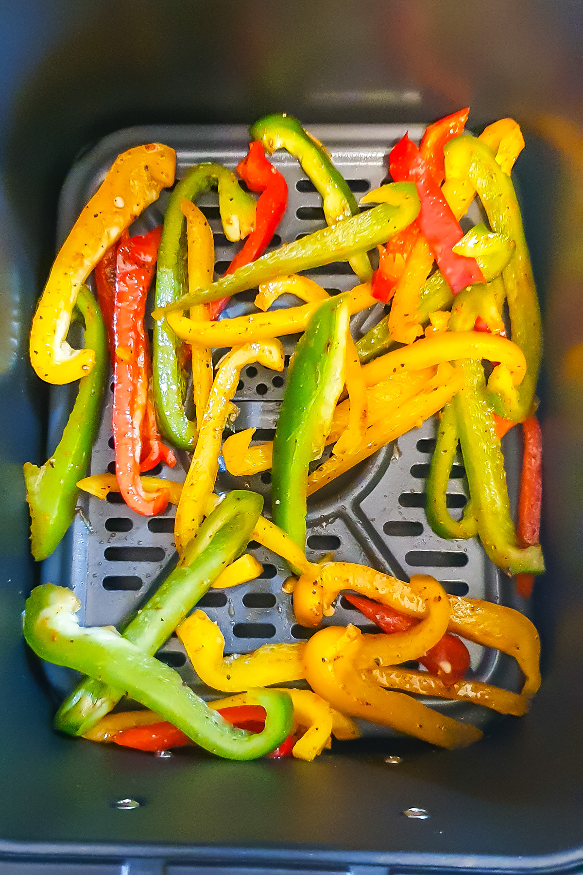Sliced green, yellow, and red bell peppers are seasoned and placed inside an air fryer basket.