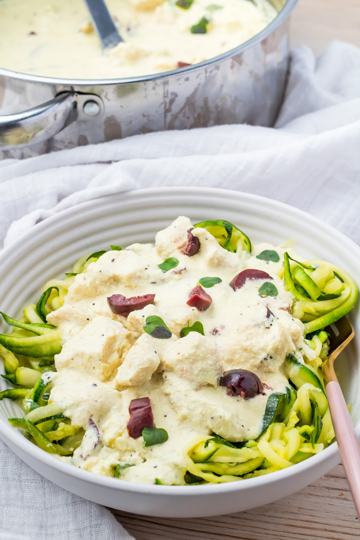 A bowl of zucchini noodles topped with creamy Greek yogurt sauce, chicken pieces, and sliced olives, with a saucepan of sauce in the background.