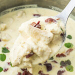 A spoonful of creamy chicken in Greek yogurt sauce with kalamata olives and fresh herbs, held above a pot filled with the same dish.