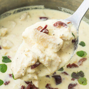 A spoonful of creamy chicken in Greek yogurt sauce with kalamata olives and fresh herbs, held above a pot filled with the same dish.