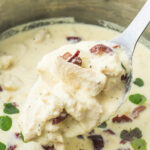 A metal ladle scoops creamy chicken in Greek yogurt sauce with kalamata olives from a stainless steel pot.