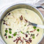 A stainless steel pot filled with chicken in greek yogurt sauce garnished with fresh herbs and olives, with a metal ladle inside.