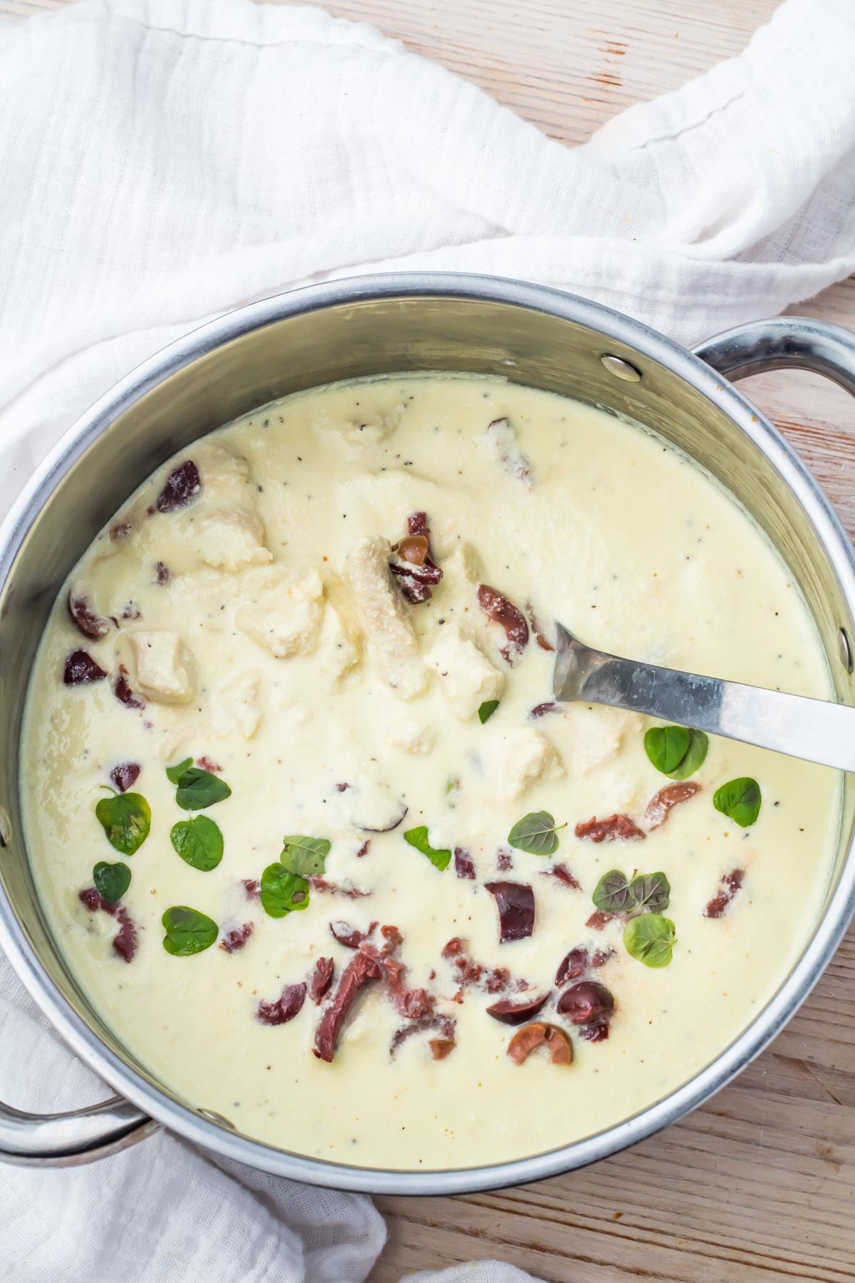 A stainless steel pot filled with chicken in greek yogurt sauce garnished with fresh herbs and olives, with a metal ladle inside.