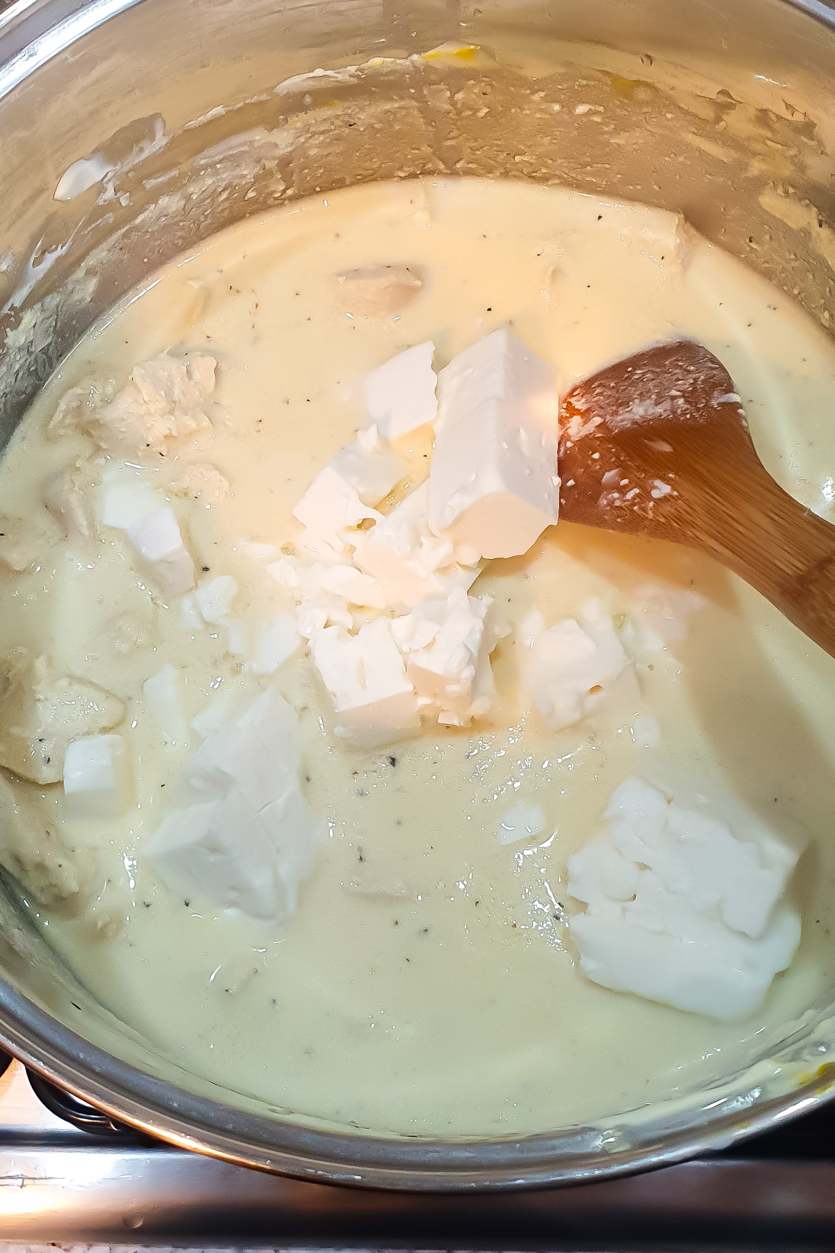 A pot of chicken in greek yogurt sauce with chunks of feta cheese being stirred with a wooden spoon.