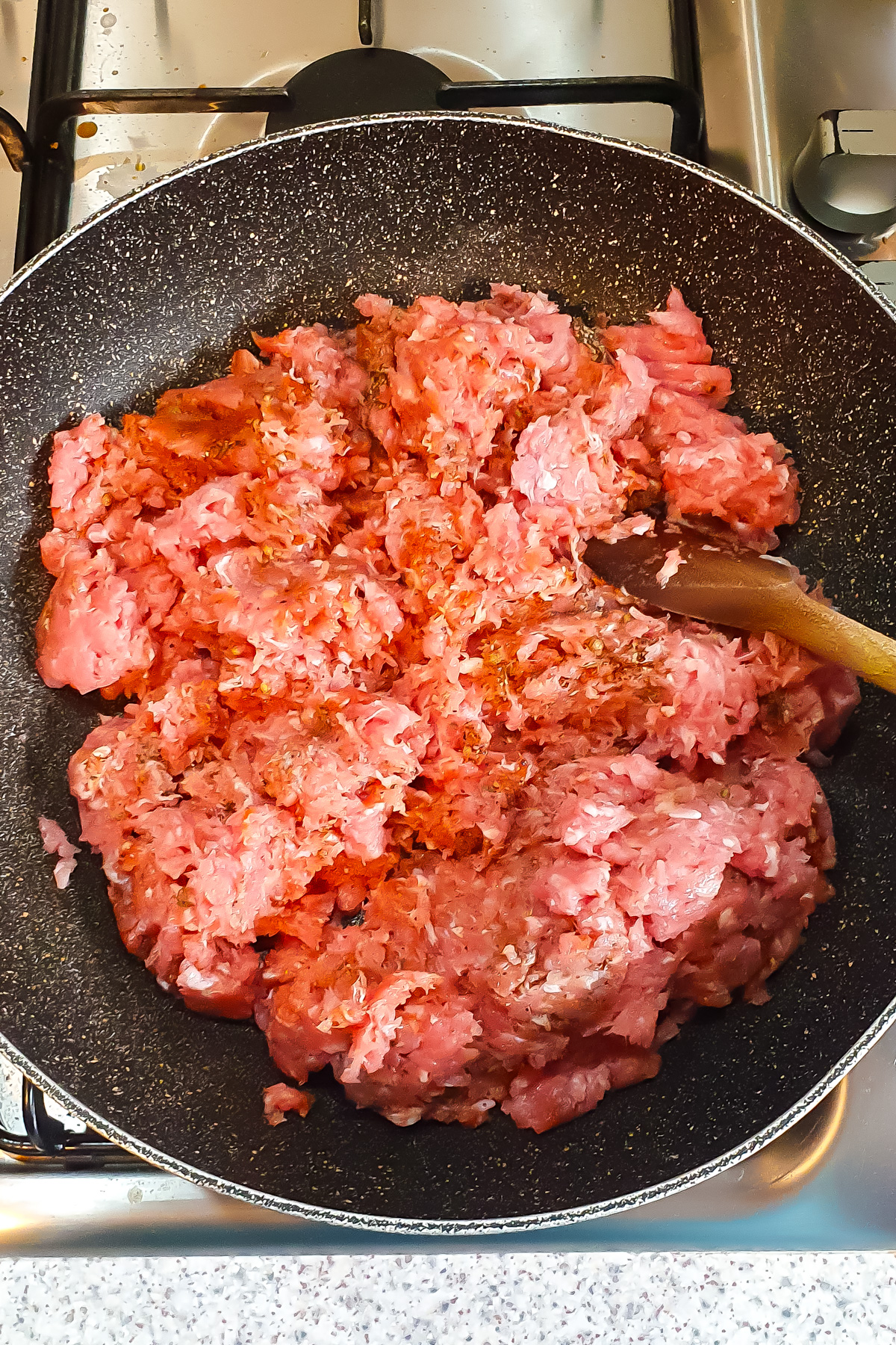 Ground meat with seasonings is being cooked in a frying pan on a stove, with a wooden spoon resting on the side of the pan.