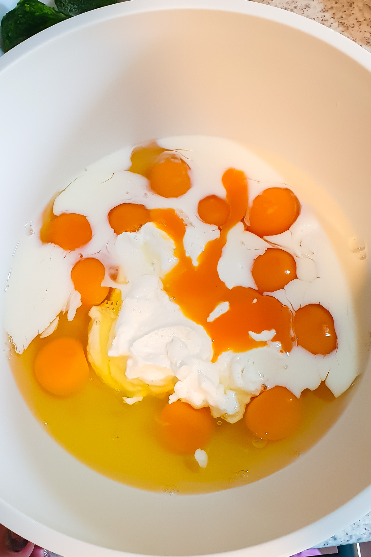 A white mixing bowl containing cracked eggs, egg yolks, liquid cream, and a dollop of white yogurt or sour cream, ready to be mixed.