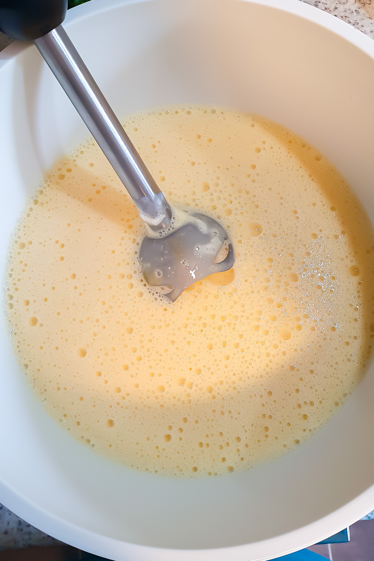 A hand blender mixing a yellow, foamy liquid in a large white mixing bowl.