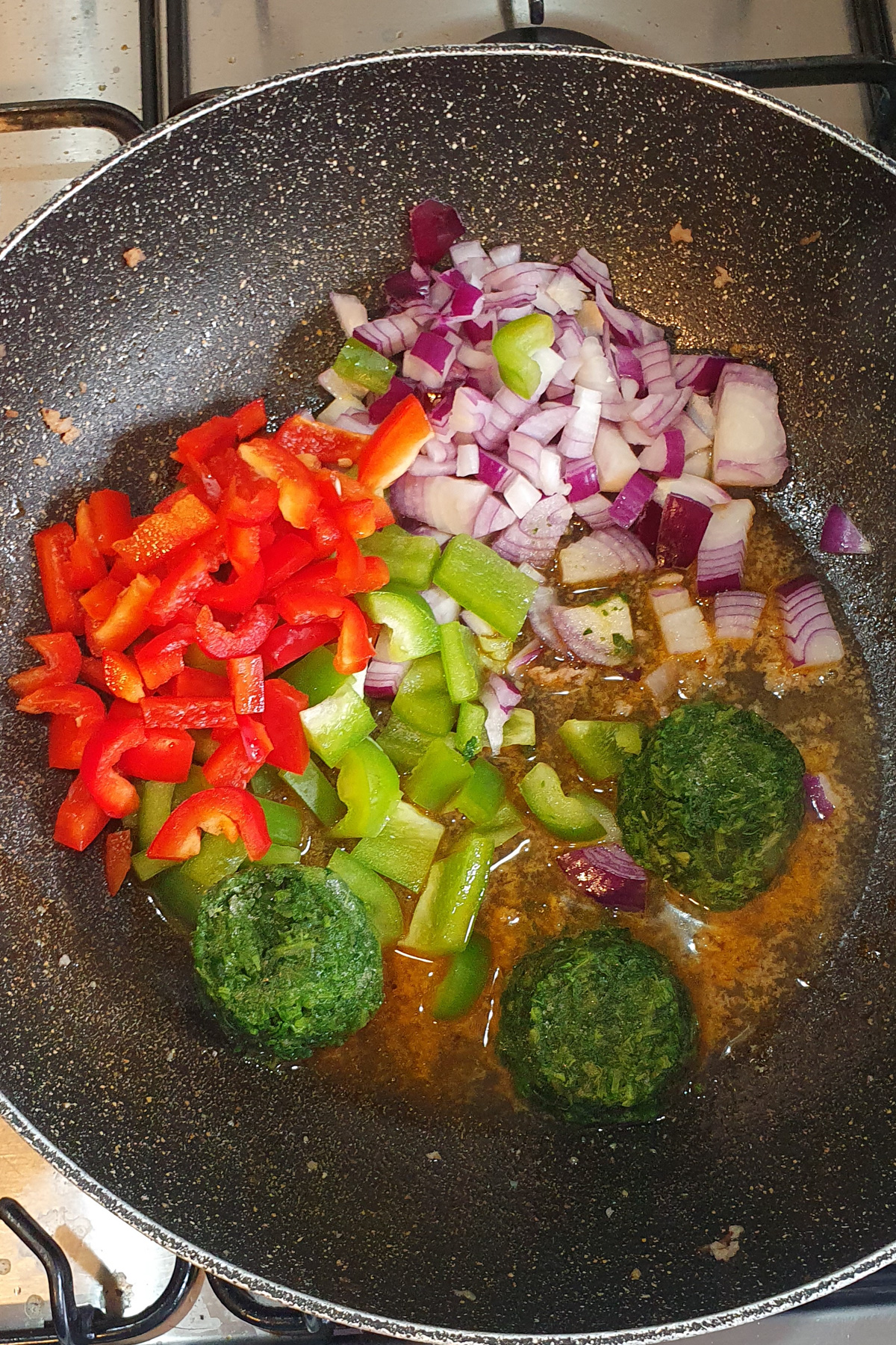 Chopped red onion, red bell pepper, green bell pepper, and three frozen spinach portions cooking in oil in a frying pan on a stove.