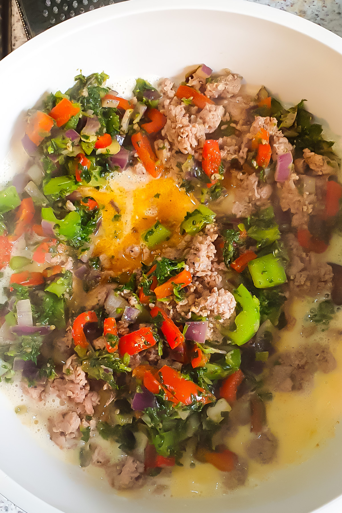 A white bowl contains raw eggs, ground meat, chopped red and green bell peppers, red onion, and leafy greens, all partially mixed together.