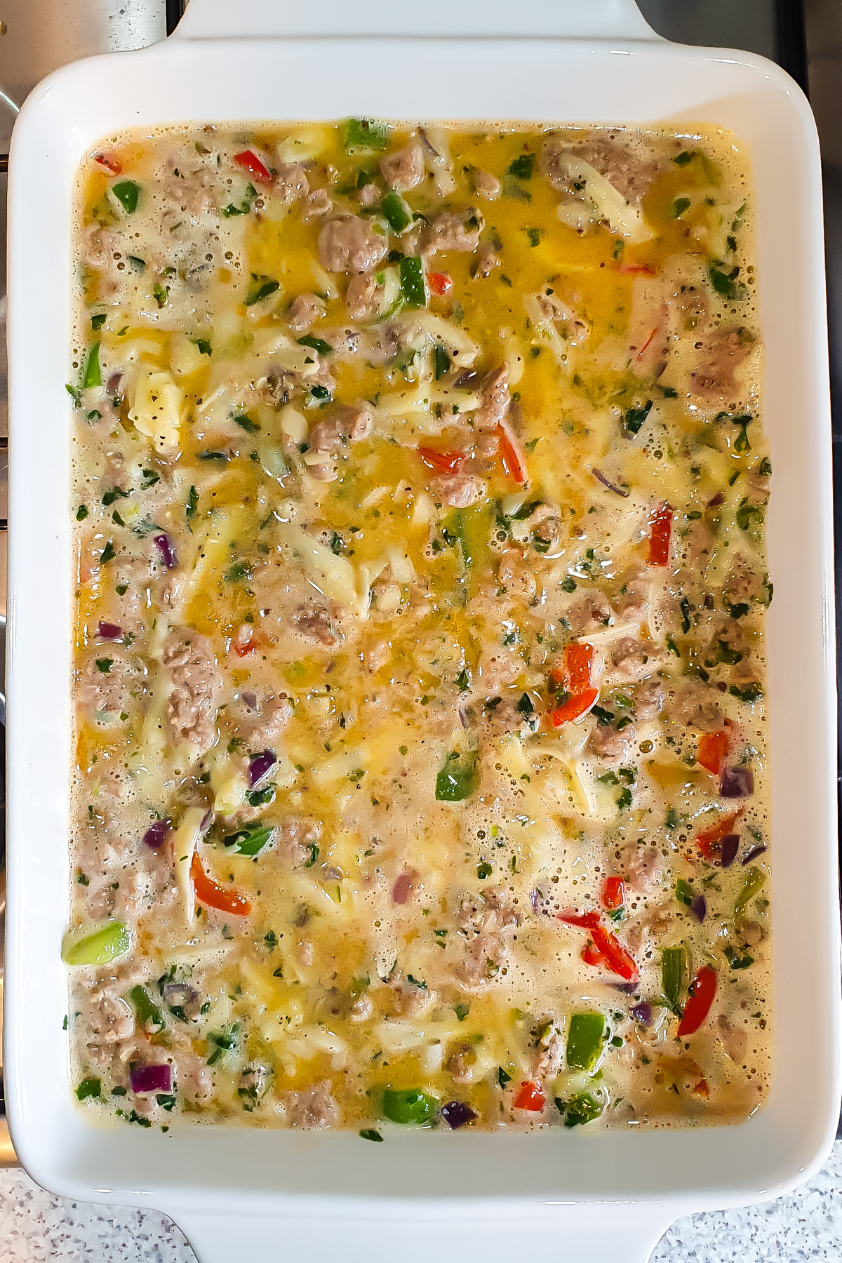 Unbaked breakfast casserole mixture with eggs, sausage, cheese, bell peppers, and onions in a white rectangular baking dish.