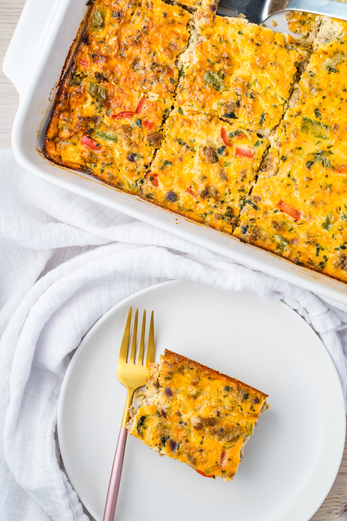 A baked sausage, egg, and cheese casserole cut into squares in a white dish, with one piece served on a white plate beside a gold fork.